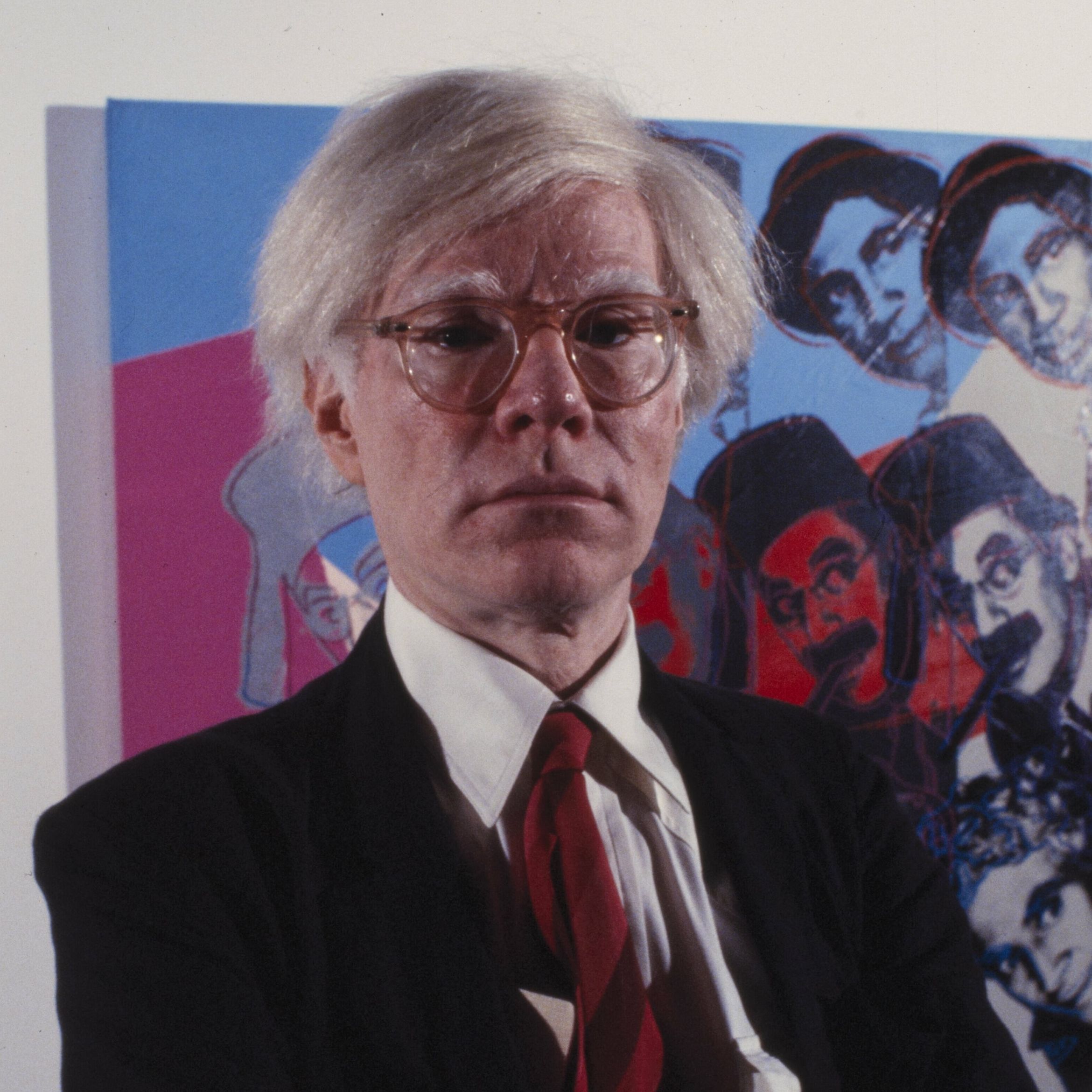 A man with white hair and glasses wearing a red tie, standing in front of a colorful artwork featuring multiple faces.
