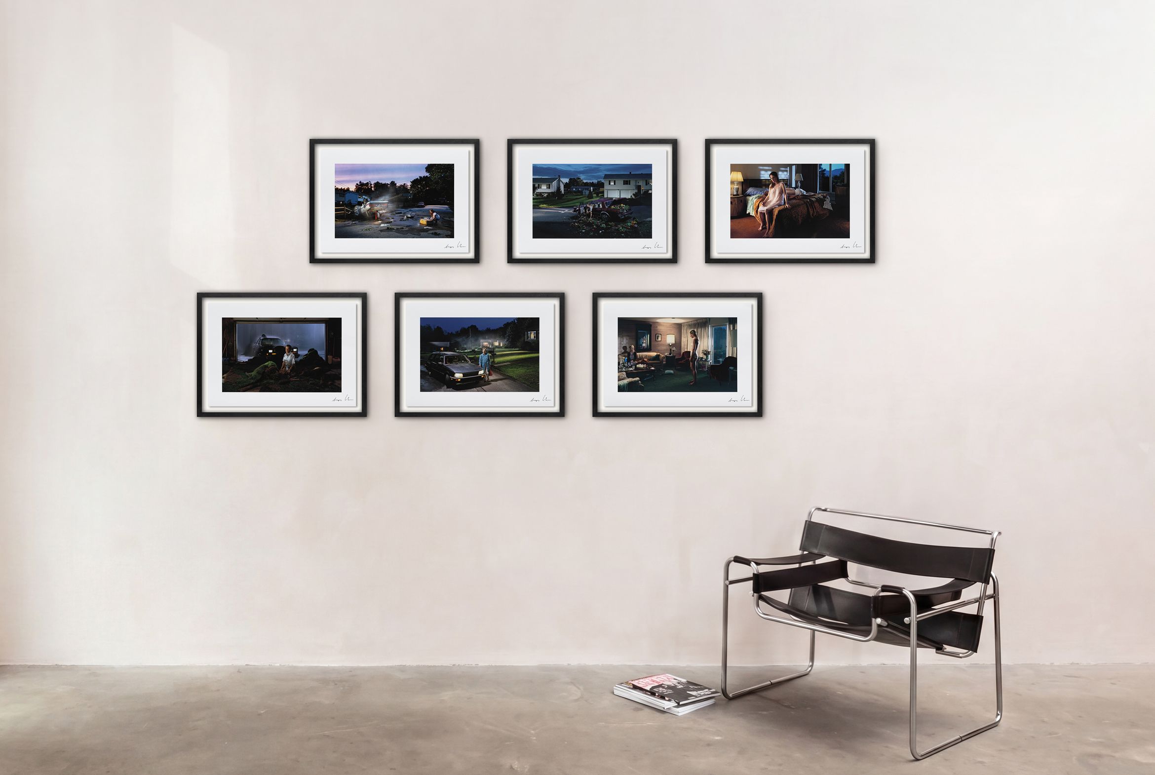 Six framed photos on a white wall above a black modern chair, with magazines on the floor nearby.