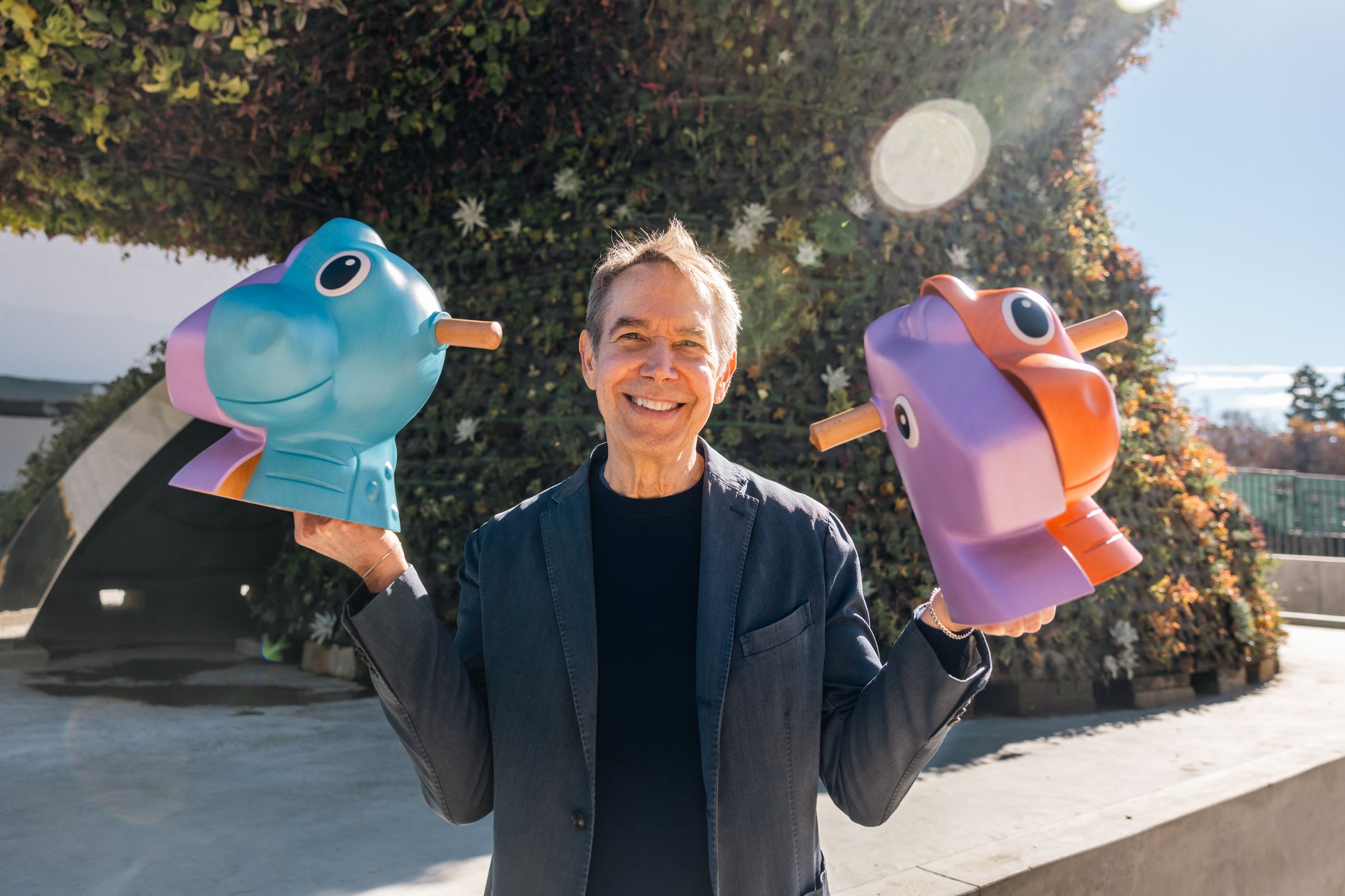 Man smiling and holding two cartoonish animal sculptures in pastel colors, standing in front of a greenery-covered structure.