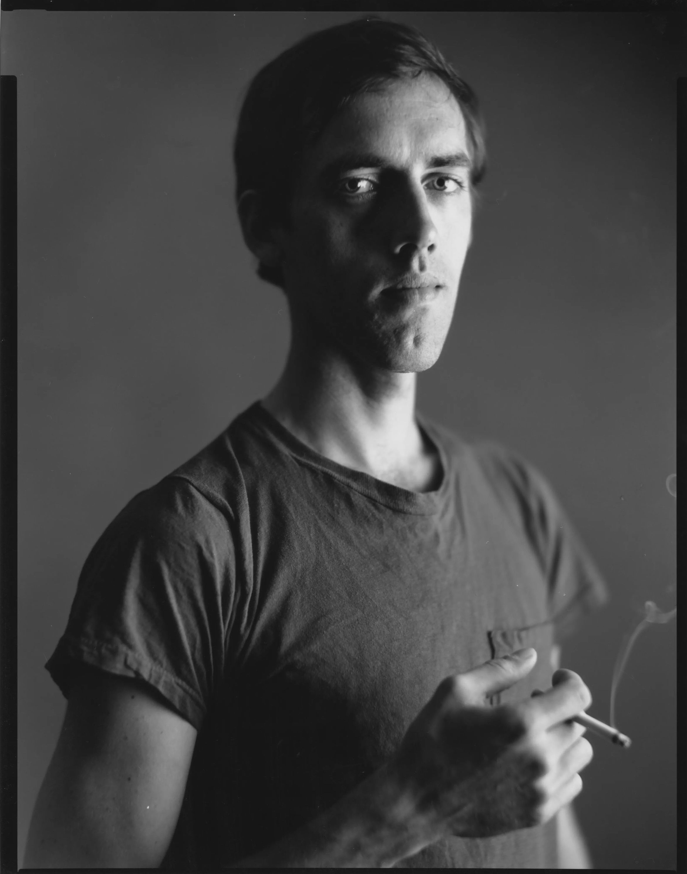 A black and white portrait of a man in a T-shirt holding a cigarette with a serious expression against a blurred background.