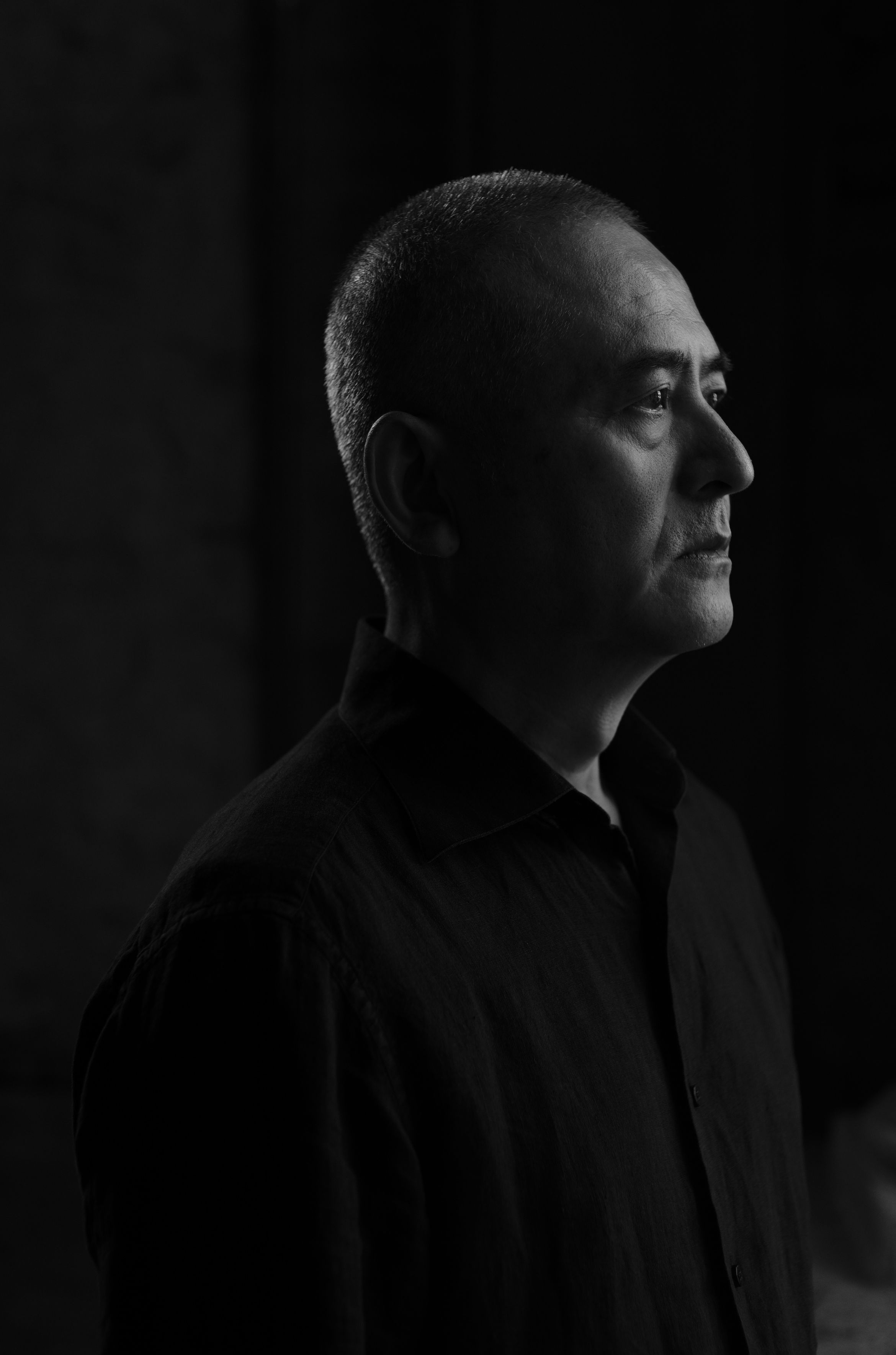 Side profile of artist Zeng Fanzhi wearing a dark shirt, gazing intently, in a dramatic black and white portrait with focused lighting.