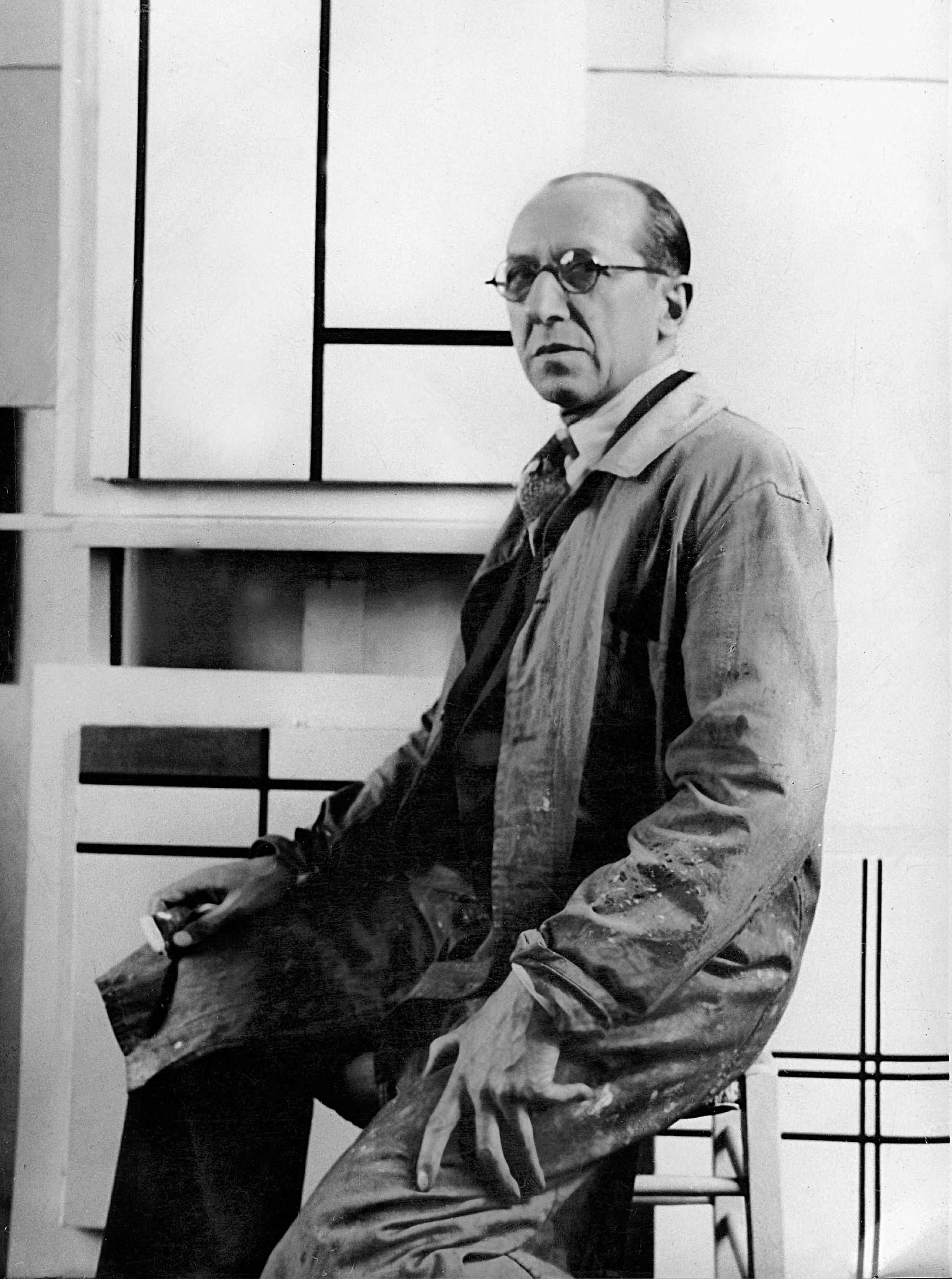 Piet Mondrian seated in front of abstract geometric paintings, wearing glasses and a paint-stained coat, looking at the camera.
