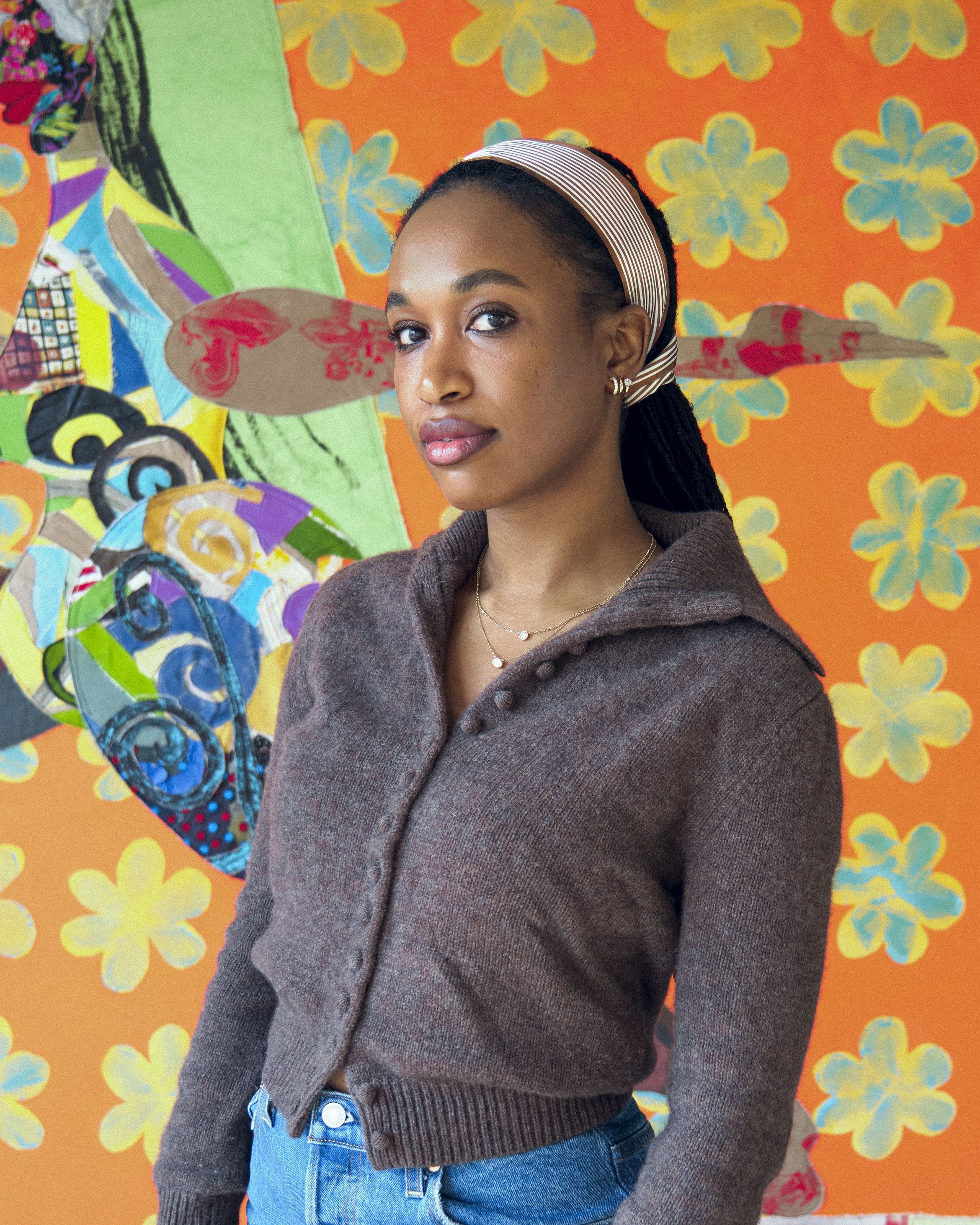 Artist Tschabalala Self in a brown sweater and jeans standing before a vibrant, orange floral and abstract-patterned background.