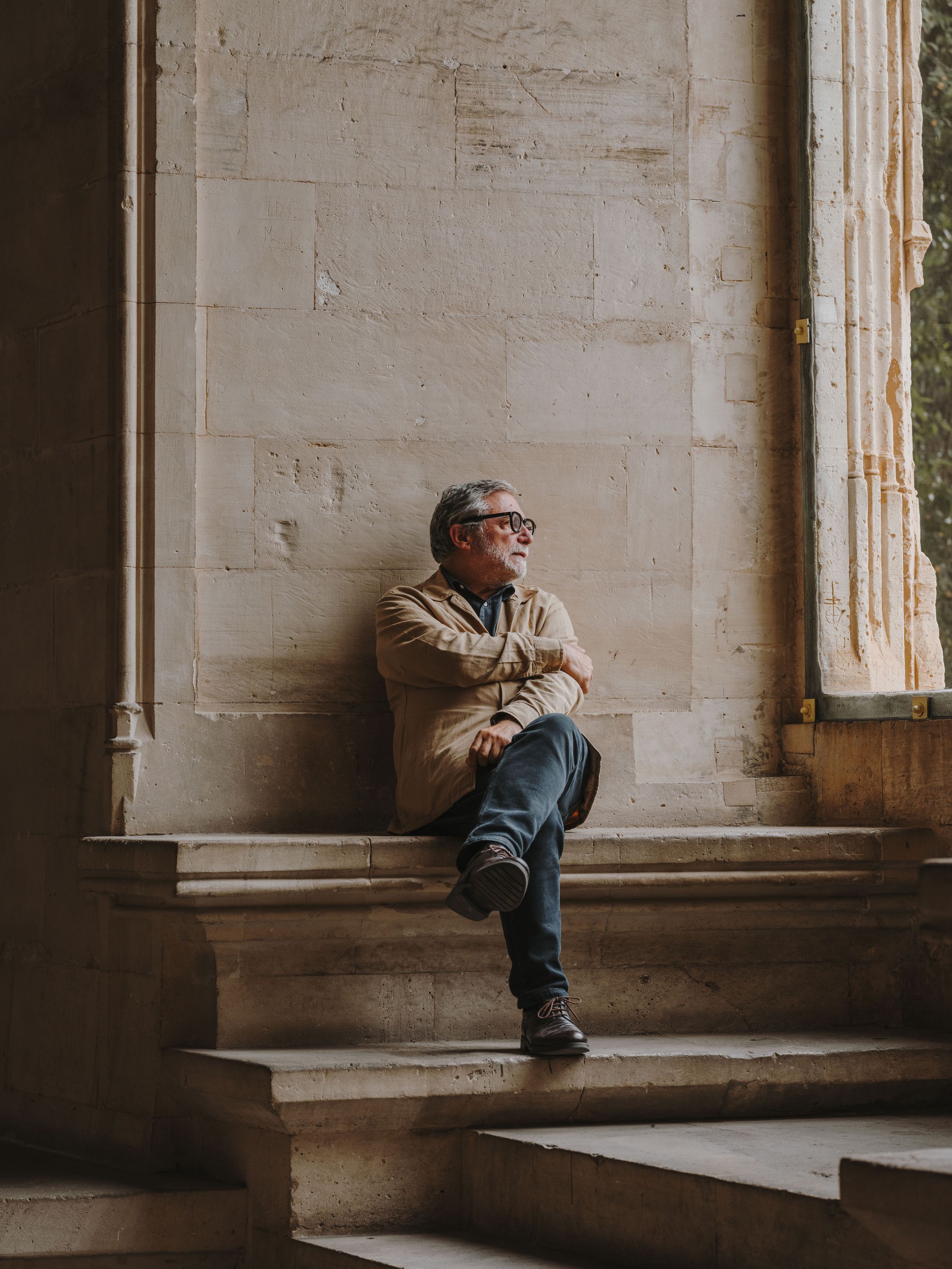 The artist Jaume Plensa in glasses and casual attire sits thoughtfully on stone steps beside a large window, gazing outside into the light.