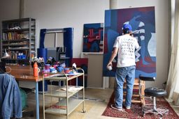 Parra's studio, with Parra at the centre, his back to the camera as he works on the large painting takes centre stage, showing a faceless blue woman in a striped dress, painted in red, purple, blue and teal. The studio is full of brightly coloured paints, with a large window on the right and a patterned rug across the floor under the painting. Parra's studio, with Parra at the centre, his back to the camera as he works on the large painting takes centre stage, showing a faceless blue woman in a striped dress, painted in red, purple, blue and teal. The studio is full of brightly coloured paints, with a large window on the right and a patterned rug across the floor under the painting.