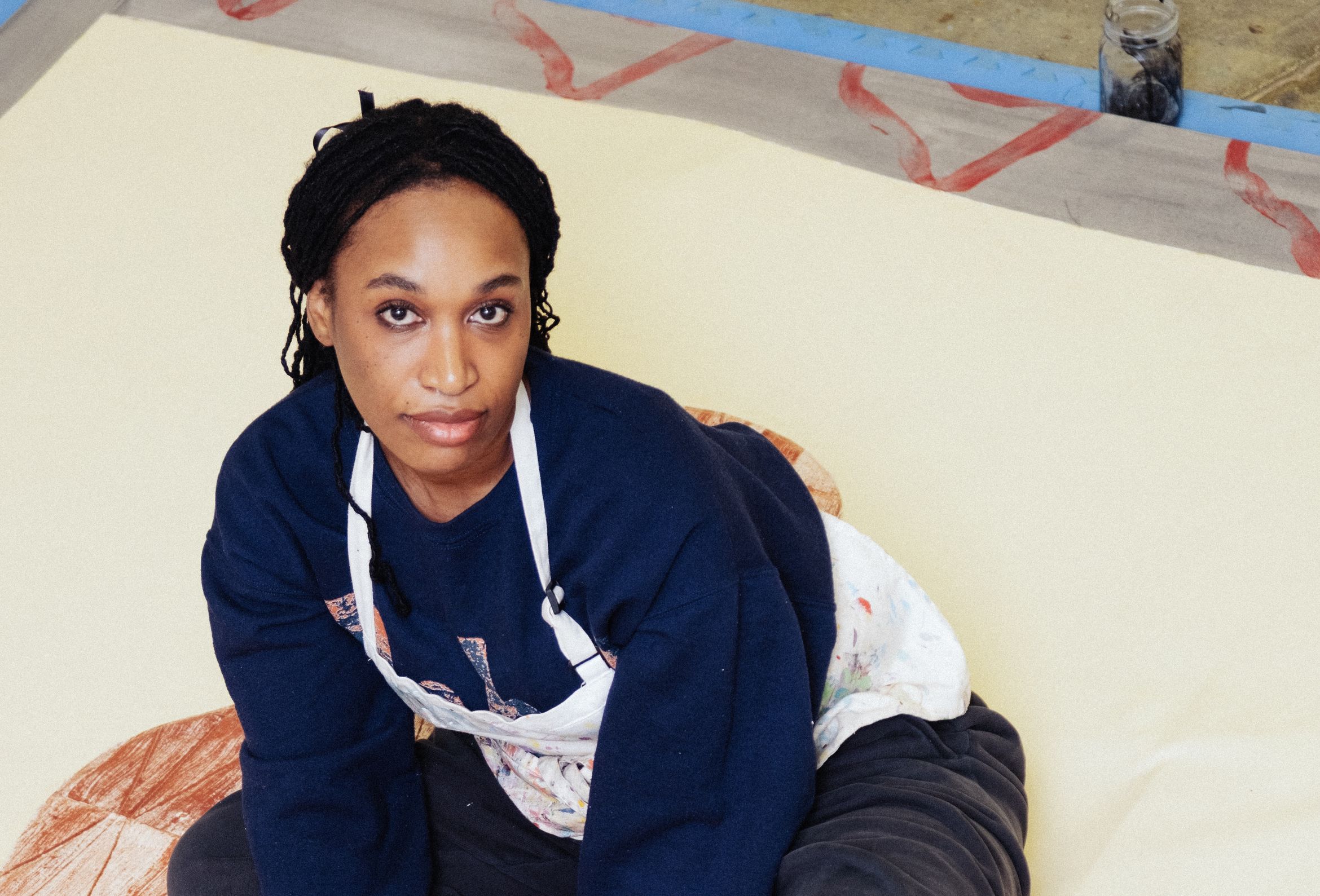 Artist sitting on a large canvas, wearing a navy sweater and paint-splattered apron, looking intently at the camera.
