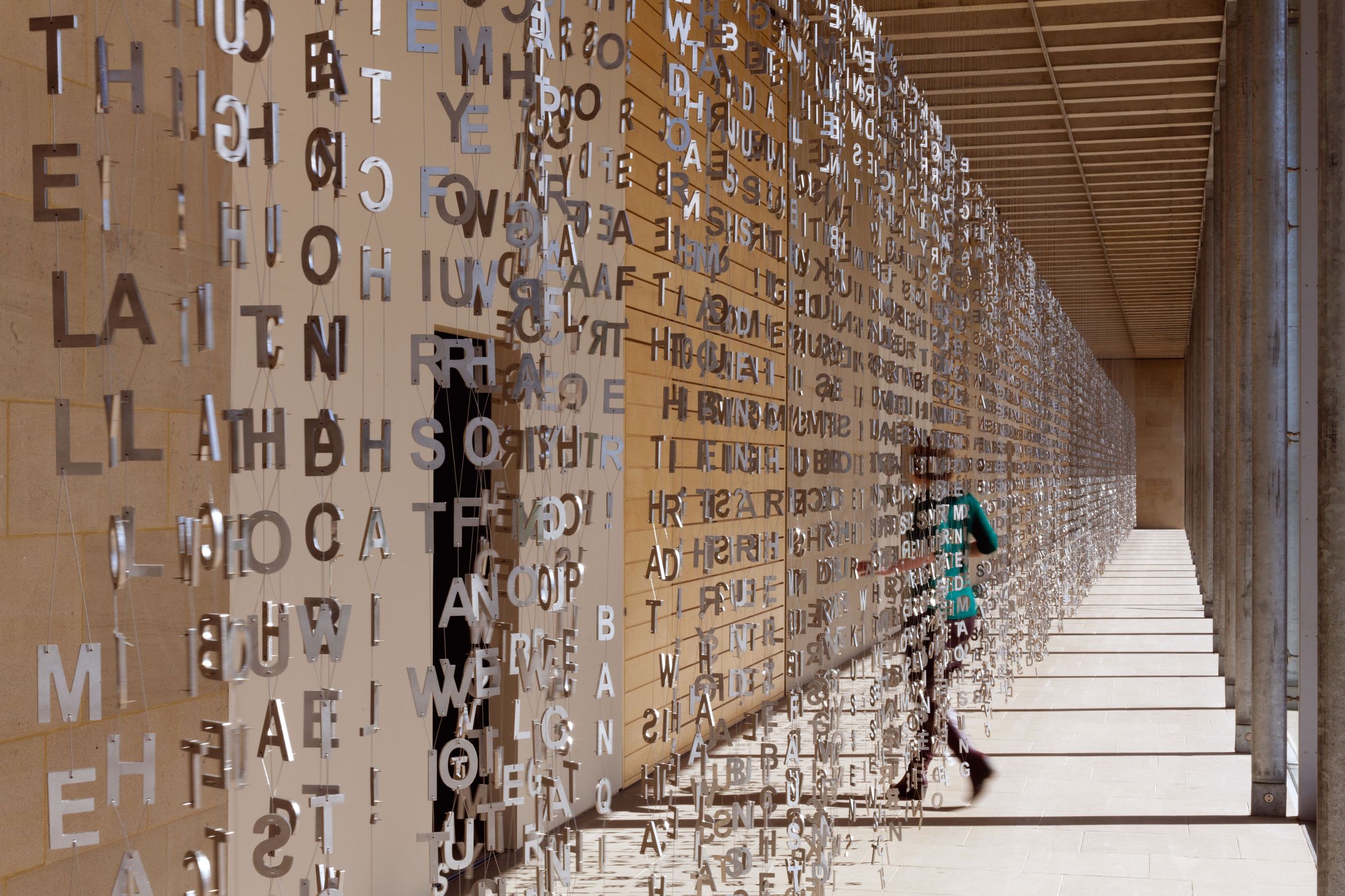 Suspended metal letters form a mesmerizing installation in a corridor, with a person walking behind them, casting shadows on the walls.
