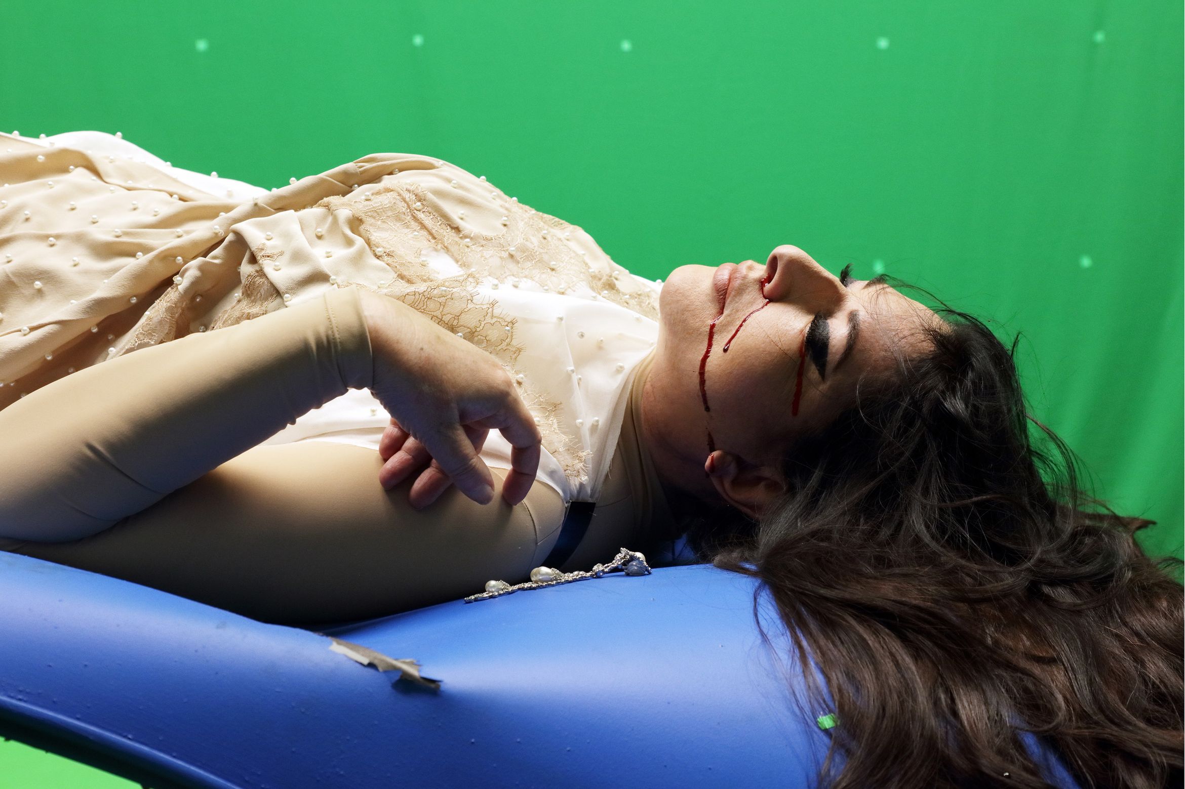 Person lying on blue cushion against green screen, wearing beaded beige gown, long dark hair, red streaks running from eyes down cheeks.