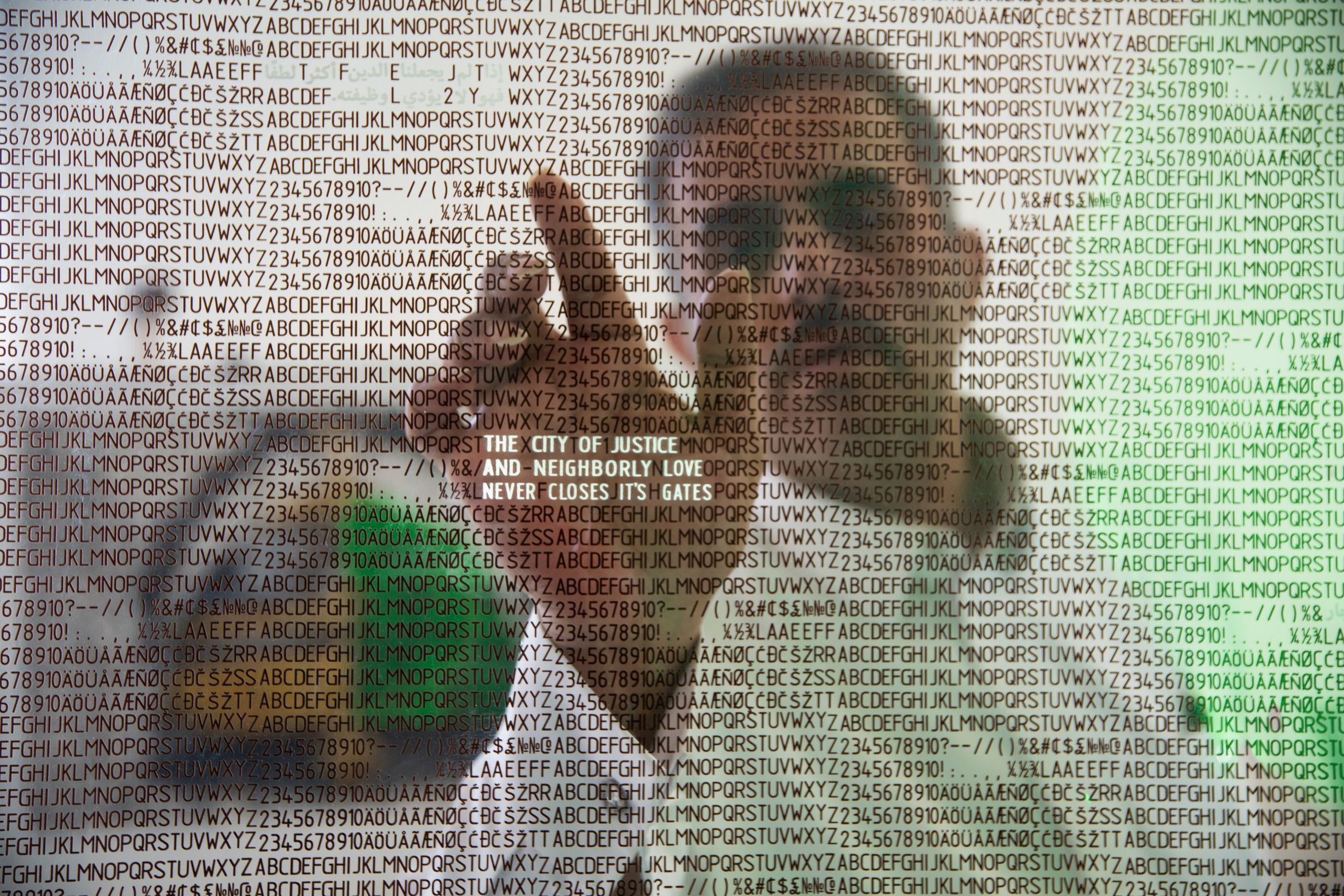 Artist Abdulnasser Gharem inspecting art - a screen of the alphabet repeating. A paragraph of text stands in contrast, reading "The city of justice and neighbourly love never closes it's gates".