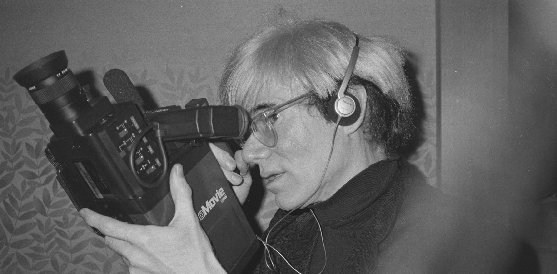 Andy Warhol with headphones using a vintage video camera, focused on capturing footage. Andy Warhol with headphones using a vintage video camera, focused on capturing footage.