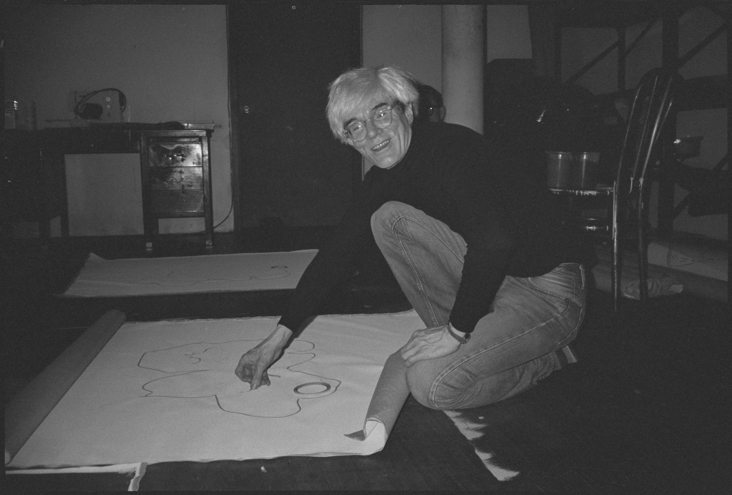 Andy Warhol, wearing a black sweater and jeans, kneels on the floor drawing on a large sheet of paper.