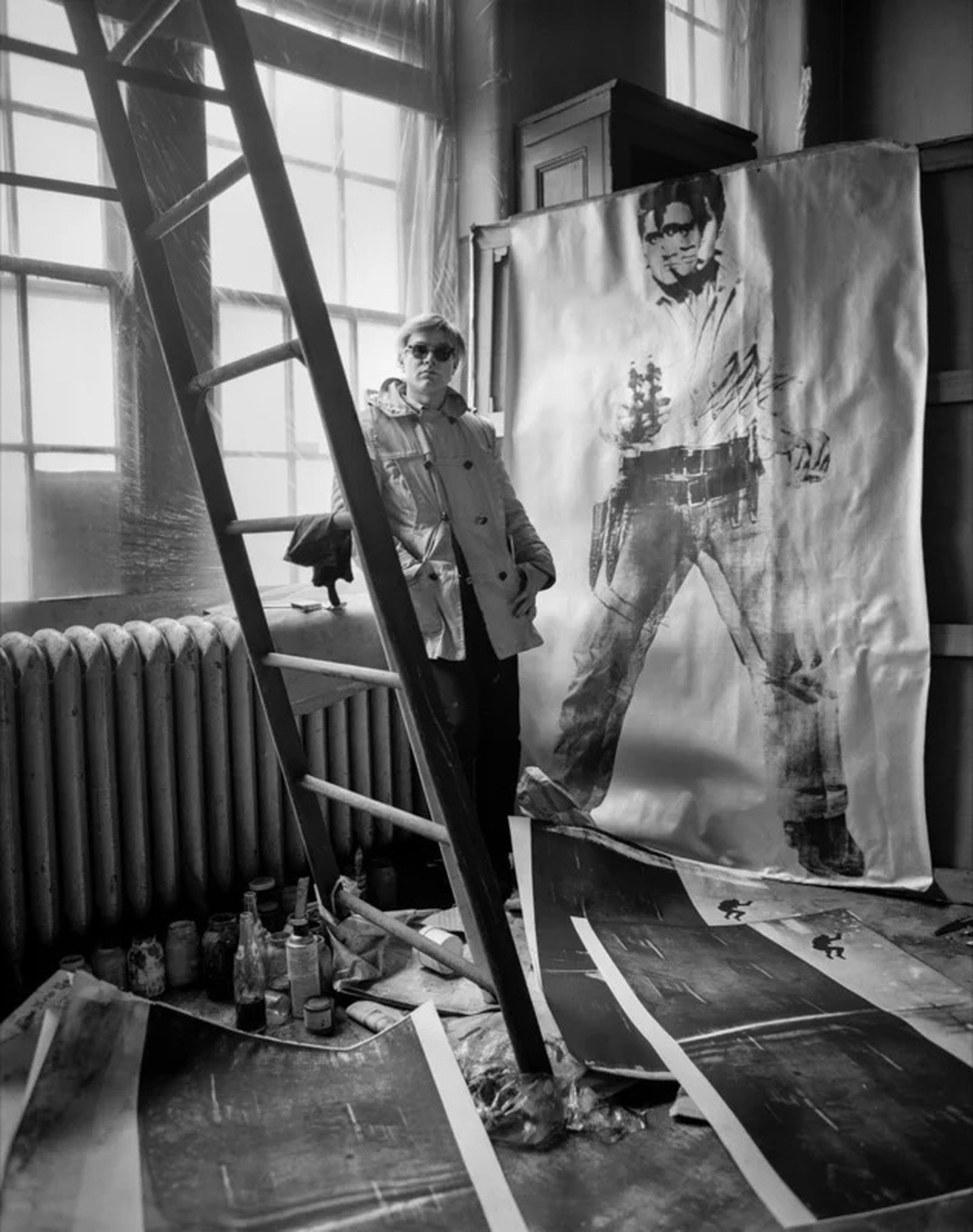 Artist Andy Warhol in sunglasses stands in an art studio next to a large print of a cowboy, surrounded by a ladder, radiator, and scattered materials.
