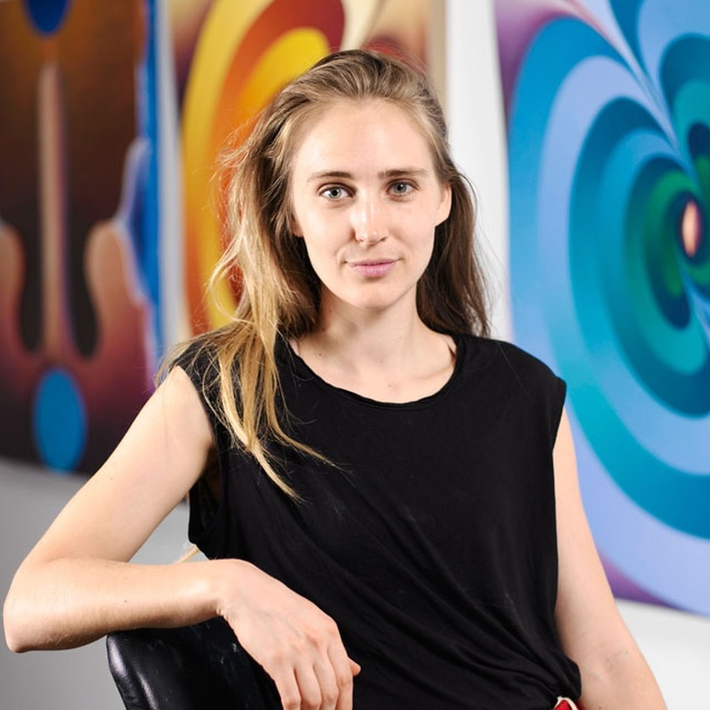 A woman in a black shirt sits in front of colorful abstract paintings, looking at the camera with a slight smile.
