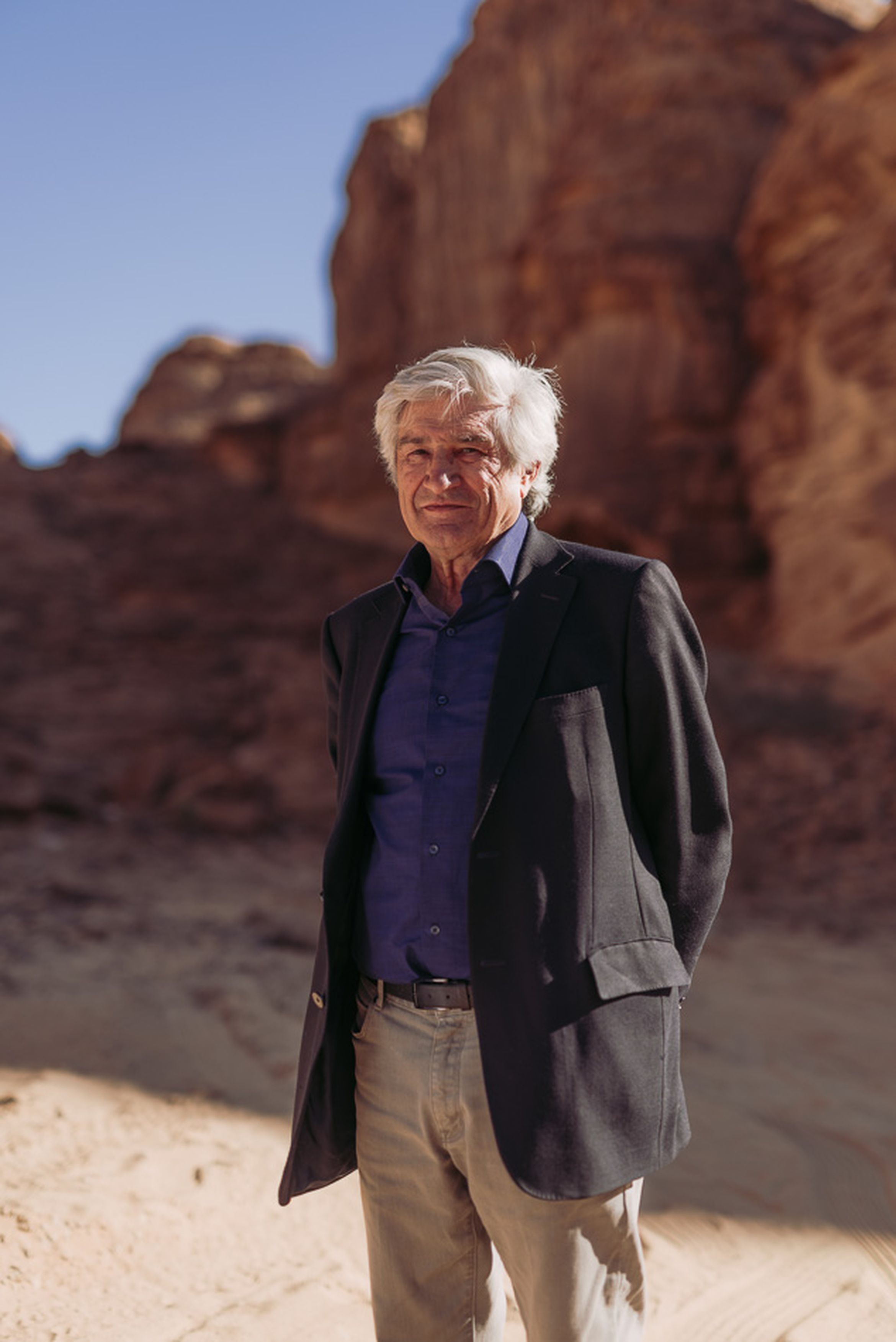 An older man in a suit standing in a sunlit desert landscape with rocky formations and a clear blue sky.