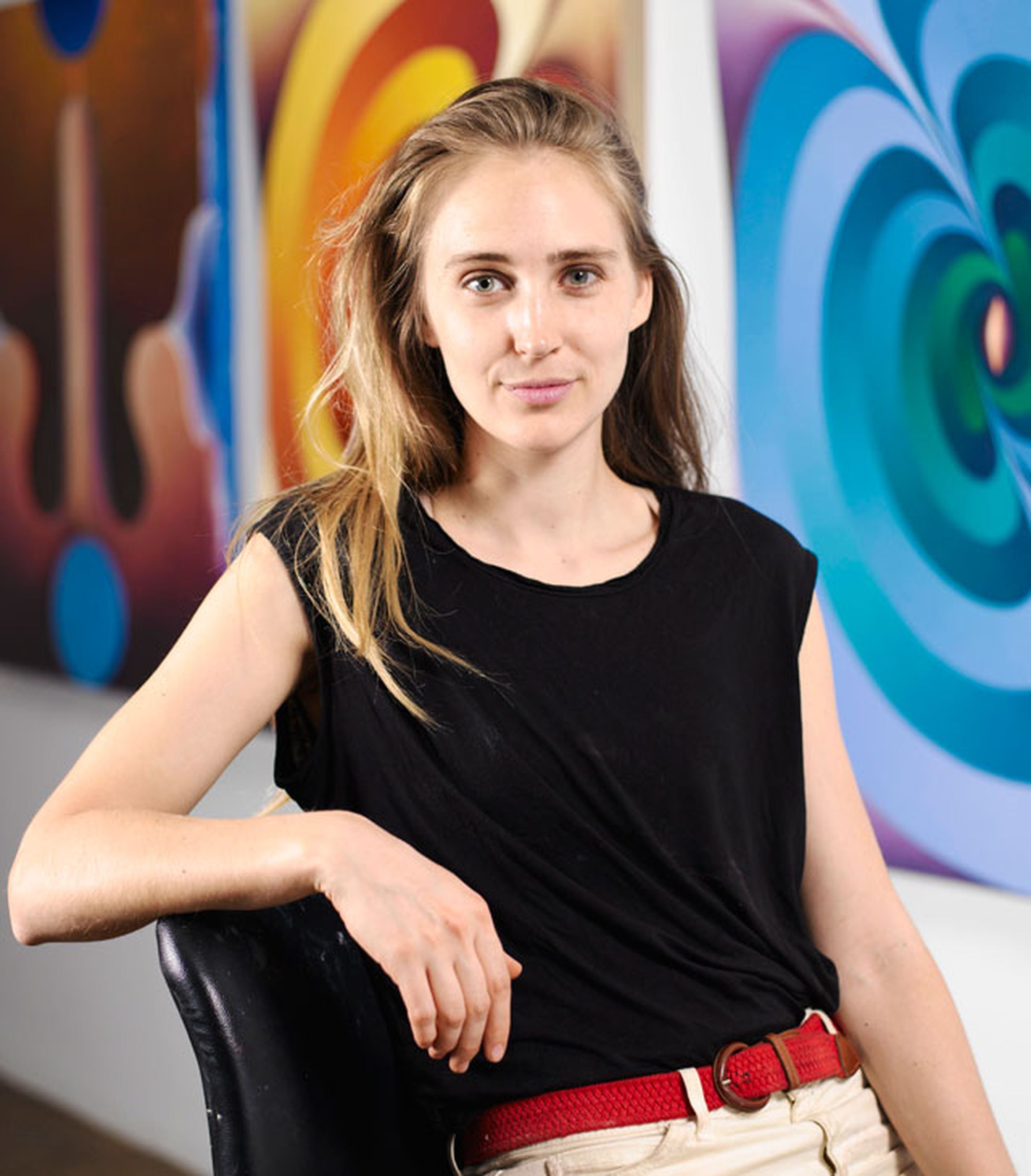 A woman with long hair in a black shirt and red belt sits confidently in front of colorful abstract paintings.