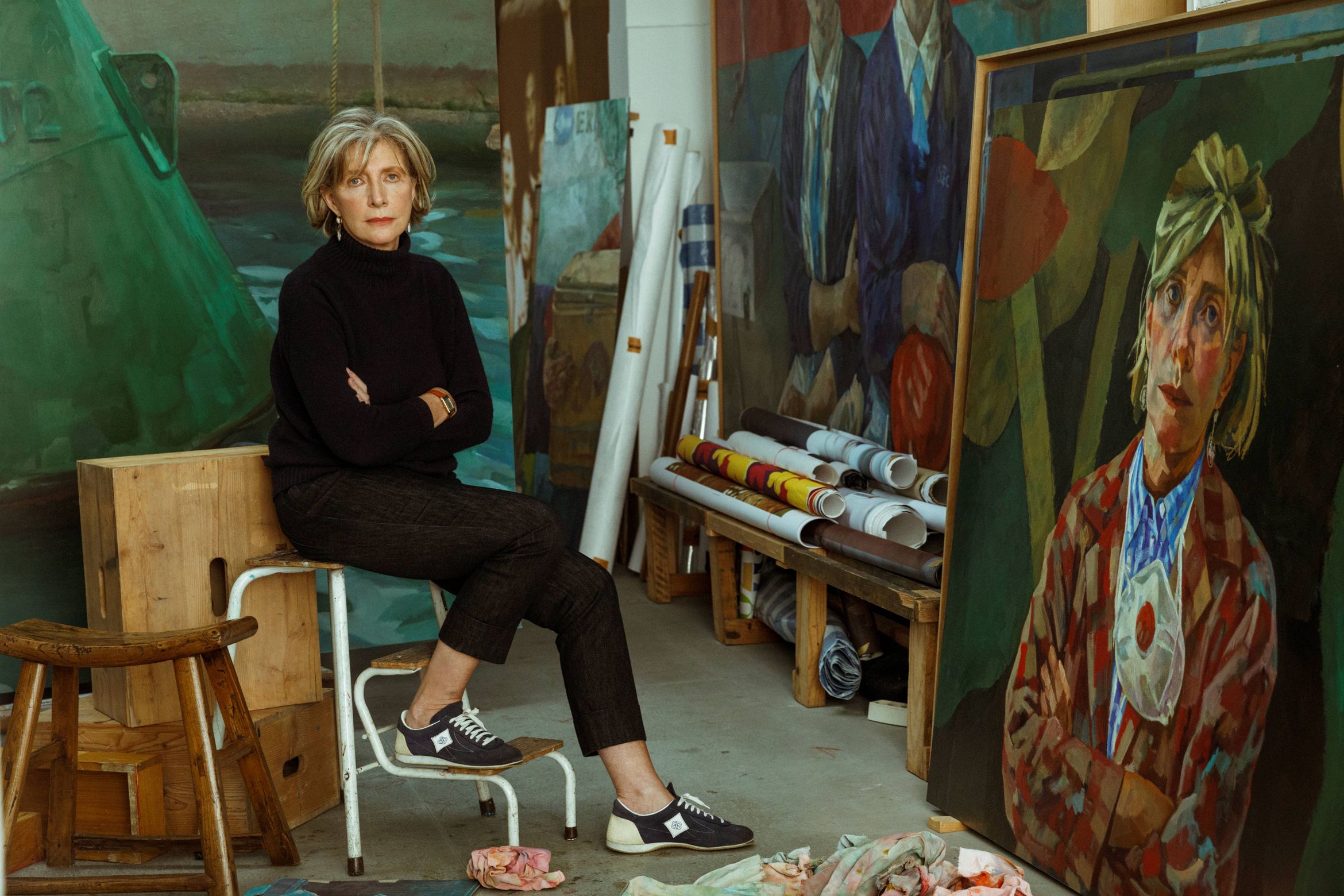 A person sitting on a stool in an art studio, surrounded by large paintings and rolled canvases, with a colorful portrait on display nearby.