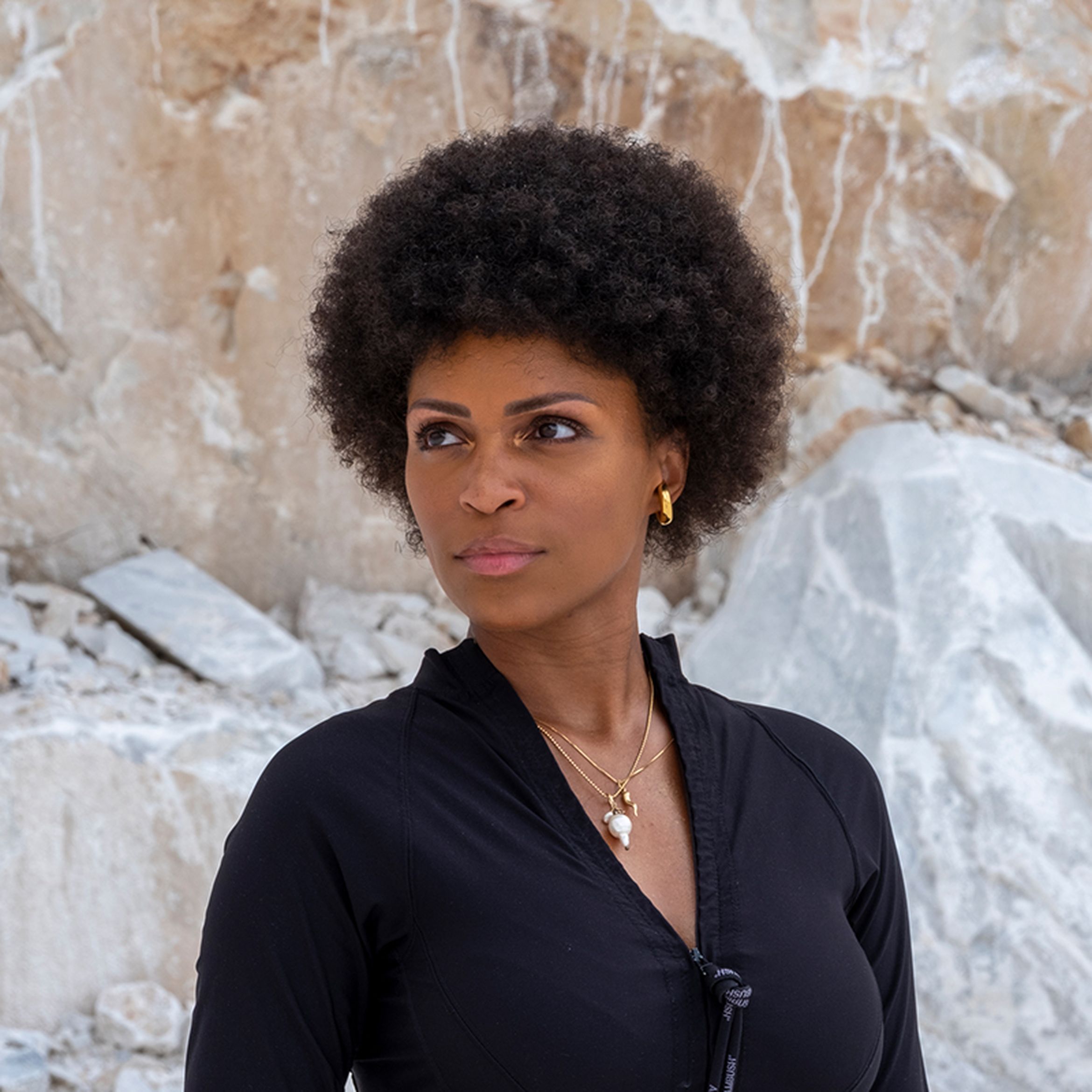 Artist wearing a black top with jewellery staring away from the camera in front of rock