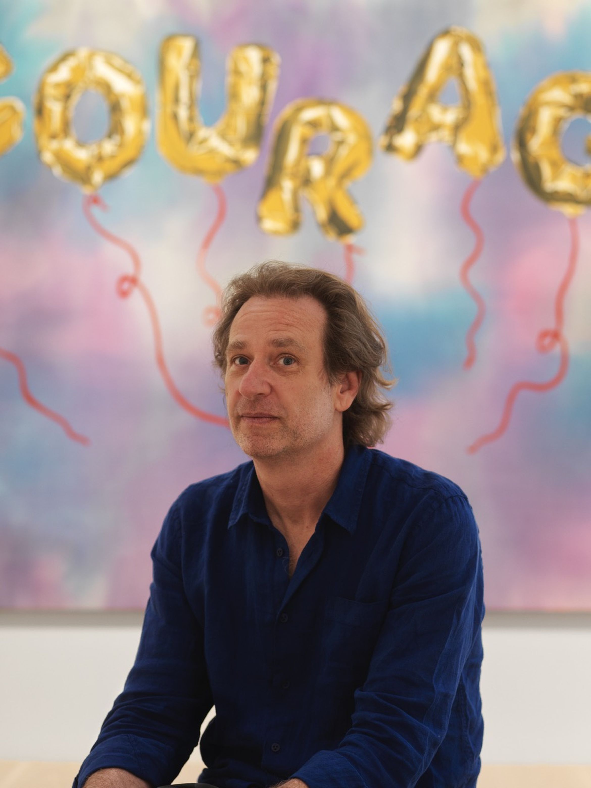 Man in blue shirt sits in front of colorful backdrop with gold balloon letters spelling "COURAGE" and red ribbon accents.