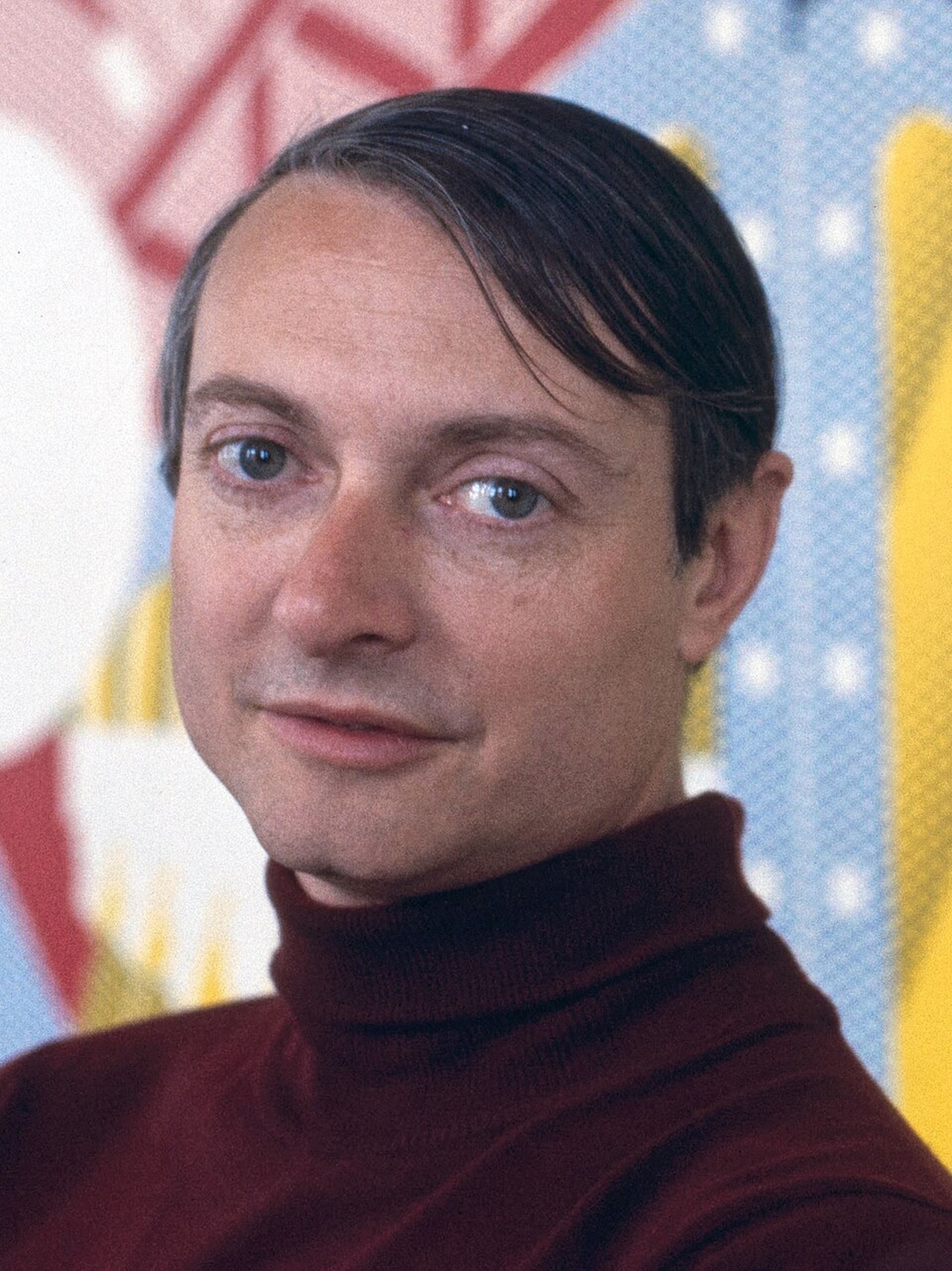 Artist Roy Lichtenstein, with short dark hair and wearing a maroon turtleneck is looking at the camera, with a colorful, abstract background.
