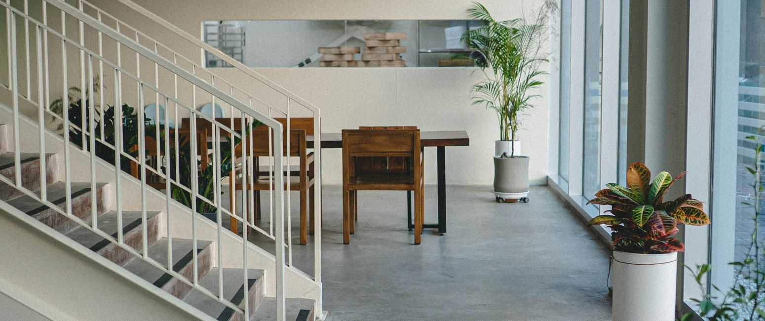 Modern interior with a staircase, wooden table and chairs, potted plants, large windows, and minimalist lighting.