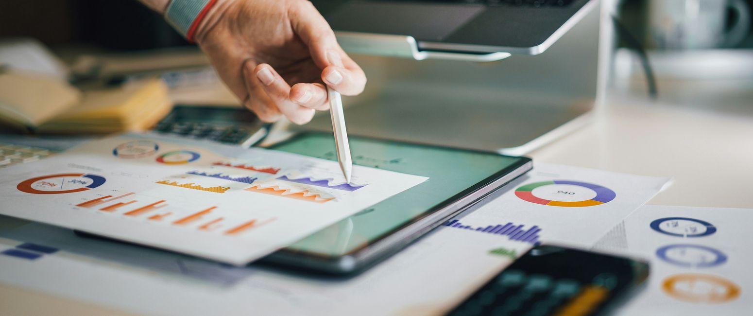 Hand pointing with pen at graphs on digital tablet, surrounded by charts, calculator, and laptop on a desk.