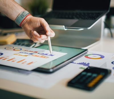 Hand pointing with pen at graphs on digital tablet, surrounded by charts, calculator, and laptop on a desk.