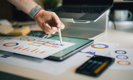 Hand pointing with pen at graphs on digital tablet, surrounded by charts, calculator, and laptop on a desk.