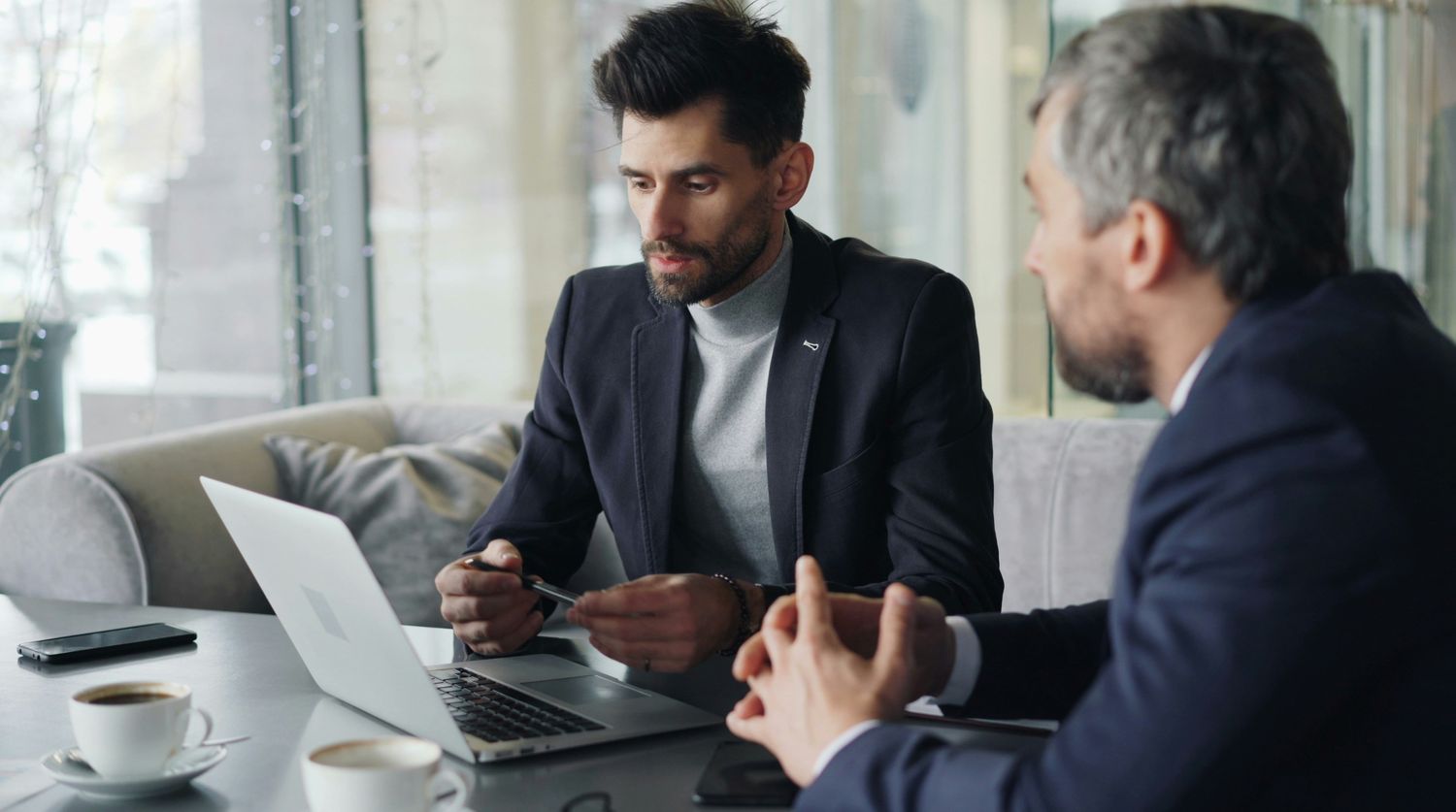 Two men in business attire engaged in discussion at a table with a laptop and coffee cups in a modern office setting.