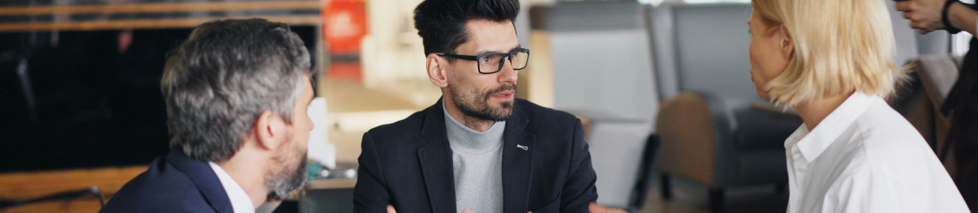 Three people in formal attire have a discussion over coffee in a modern office setting with large windows and bright lighting.