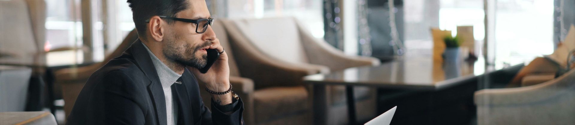Man in a suit talks on a smartphone while working on a laptop at a café table, with coffee nearby and a modern interior in the background.