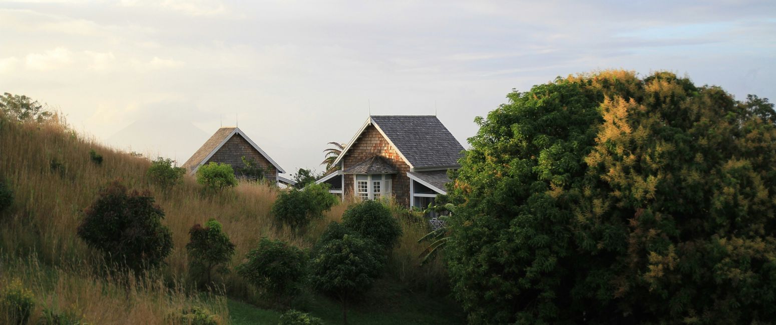 A cottage with a shingled roof is nestled between lush trees and grassy hills under a partly cloudy sky.