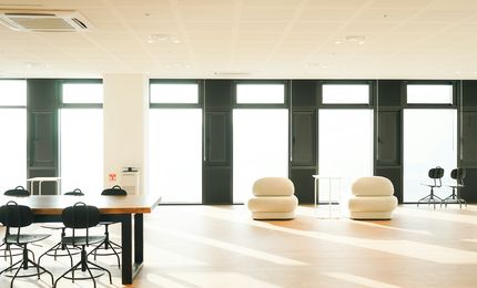Modern, spacious office lounge with large windows, natural light, minimalist furniture, and wooden flooring. Tables and chairs are neatly arranged.