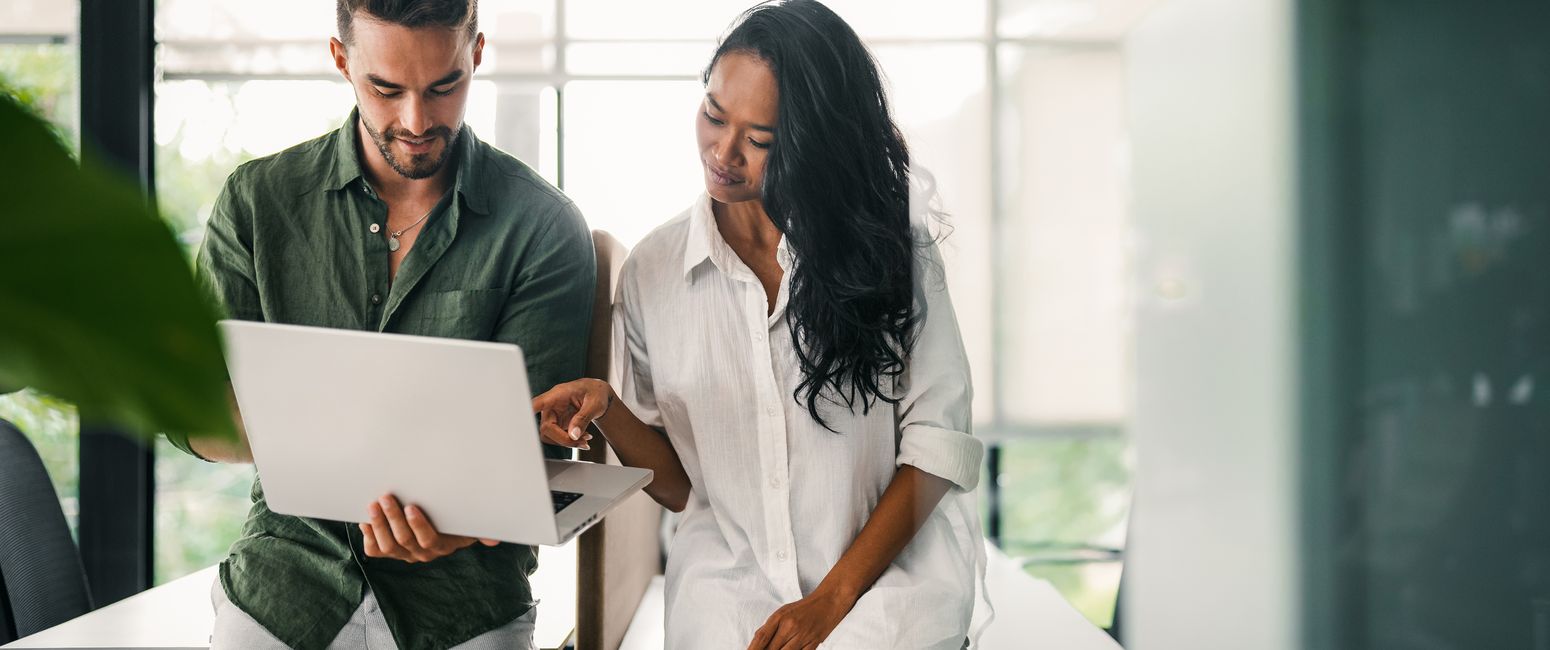 Two colleagues, a man and a woman, discuss work on a laptop while standing in a modern, well-lit office with large windows.