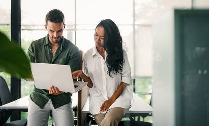 Two colleagues, a man and a woman, discuss work on a laptop while standing in a modern, well-lit office with large windows.