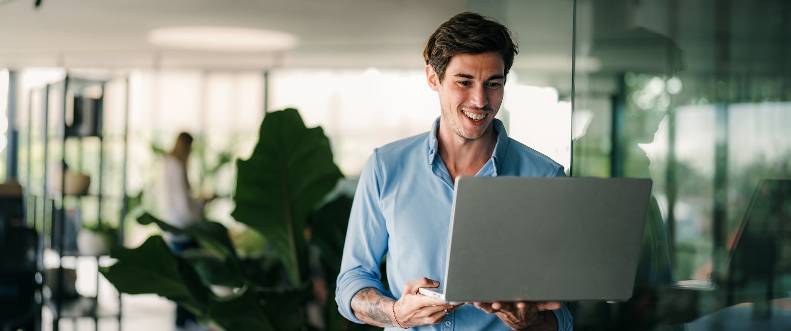 Smiling man in a blue shirt holds a laptop, standing near a glass wall in a modern office with plants in the background.