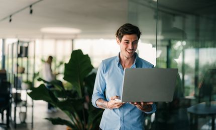 Smiling man in a blue shirt holds a laptop, standing near a glass wall in a modern office with plants in the background.
