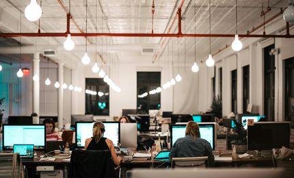 Open-plan office with people working at desks, lit by multiple hanging lights, creating a focused and collaborative atmosphere.