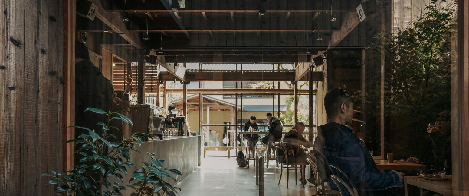 Cozy cafe interior with people seated at tables, wooden walls, greenery, and a barista behind the counter. Sunlight illuminates the space.