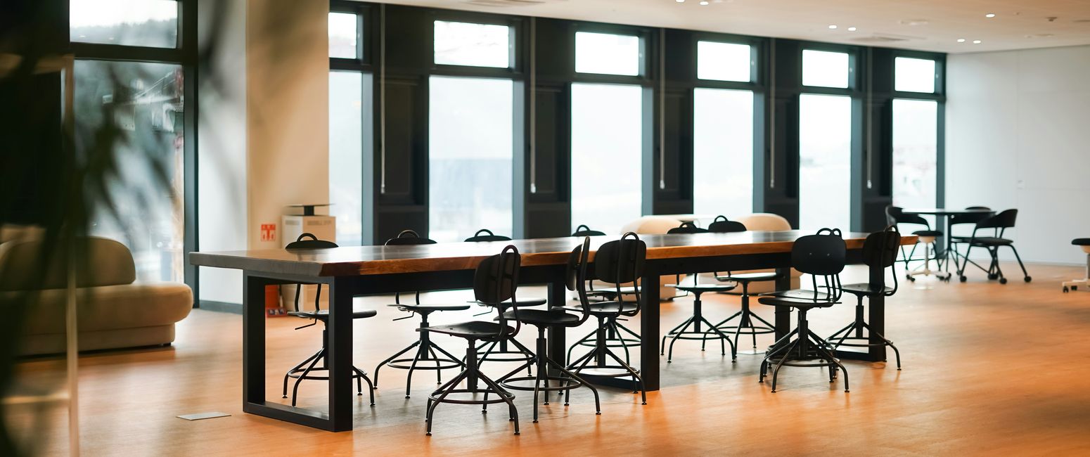 Spacious, modern office with large windows, tables, and chairs on a wooden floor, softly lit by daylight.