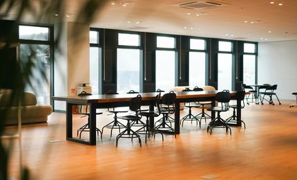 Spacious, modern office with large windows, tables, and chairs on a wooden floor, softly lit by daylight.