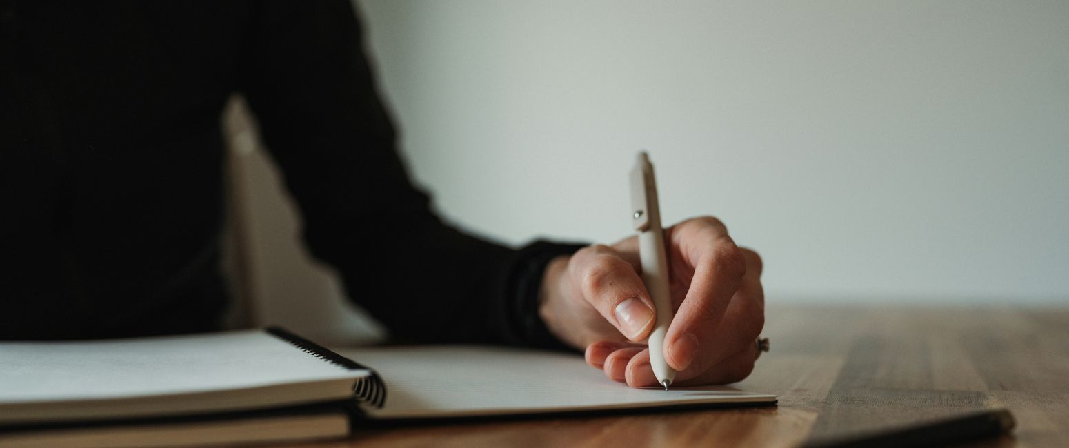 A person writes in a notebook with a pen on a wooden table. Only the hand and arm are visible, creating a focused and intimate scene.