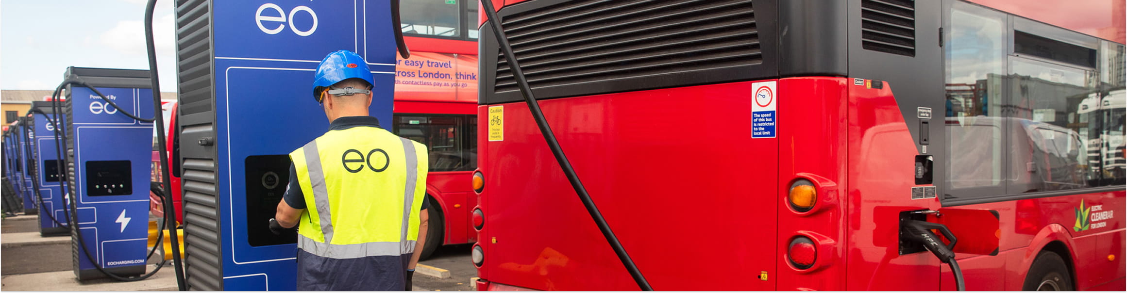 Technician in a hi-vis vest operates an electric bus charging station next to a red bus, with more buses and charging units in the background.
