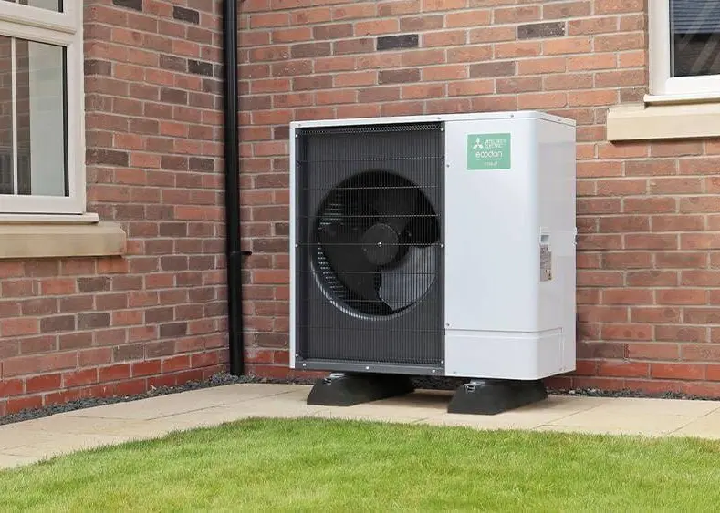 A white outdoor heat pump unit is mounted on a brick wall beside a window, positioned on a small concrete slab on a grassy area.