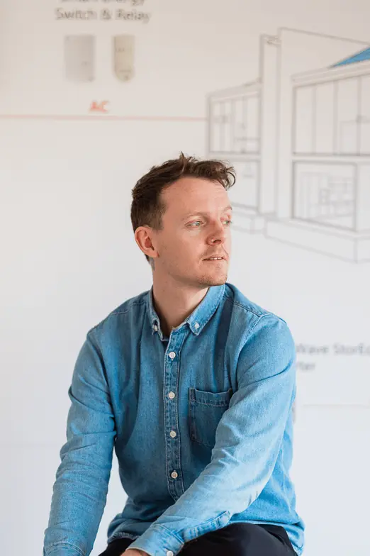 Man in a blue denim shirt sits thoughtfully in front of a blurred architectural diagram on a whiteboard.