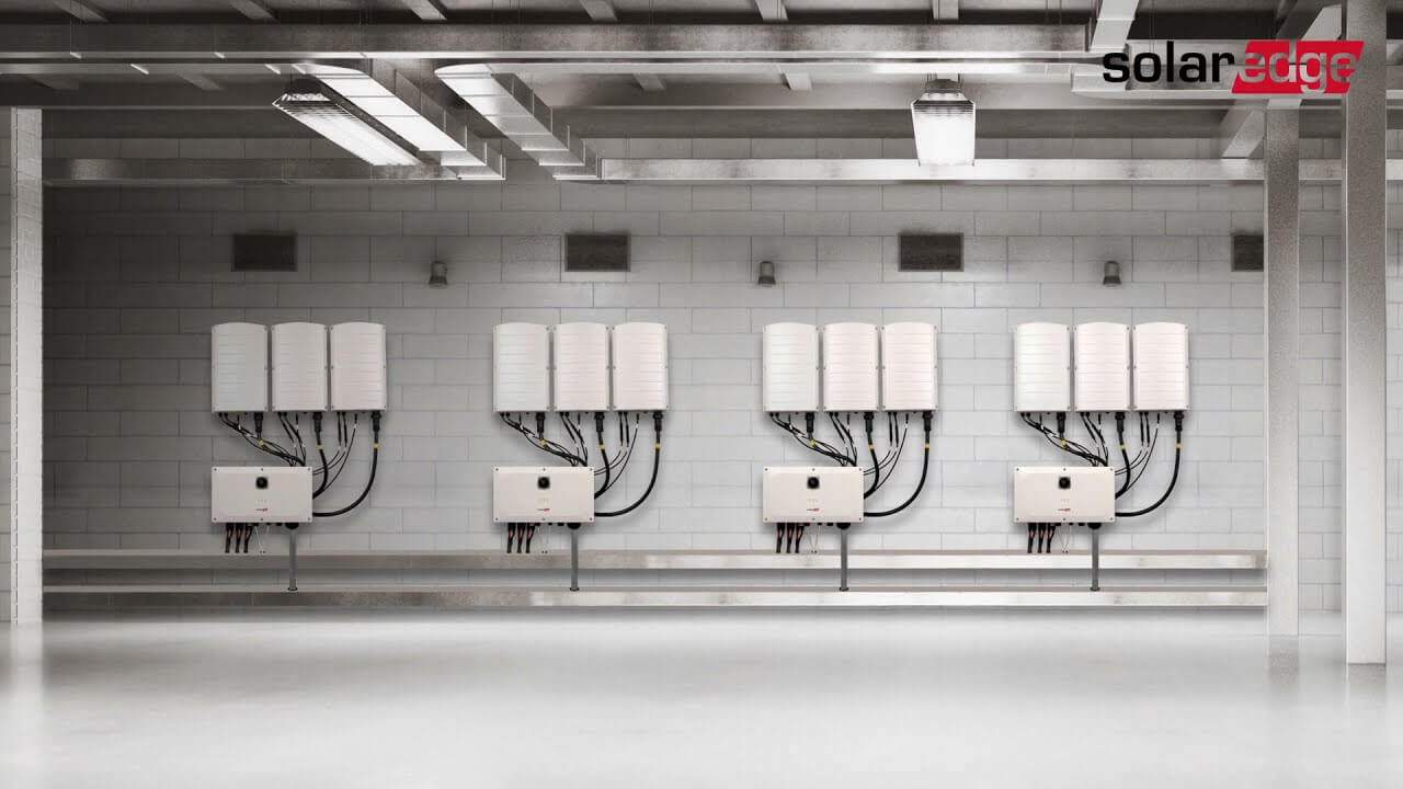 A row of solar inverters mounted on a white brick wall in an industrial setting, with cables neatly arranged below each unit.
