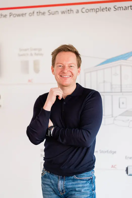 Smiling person in a navy shirt and jeans stands in front of a display about smart energy solutions, with arms crossed.