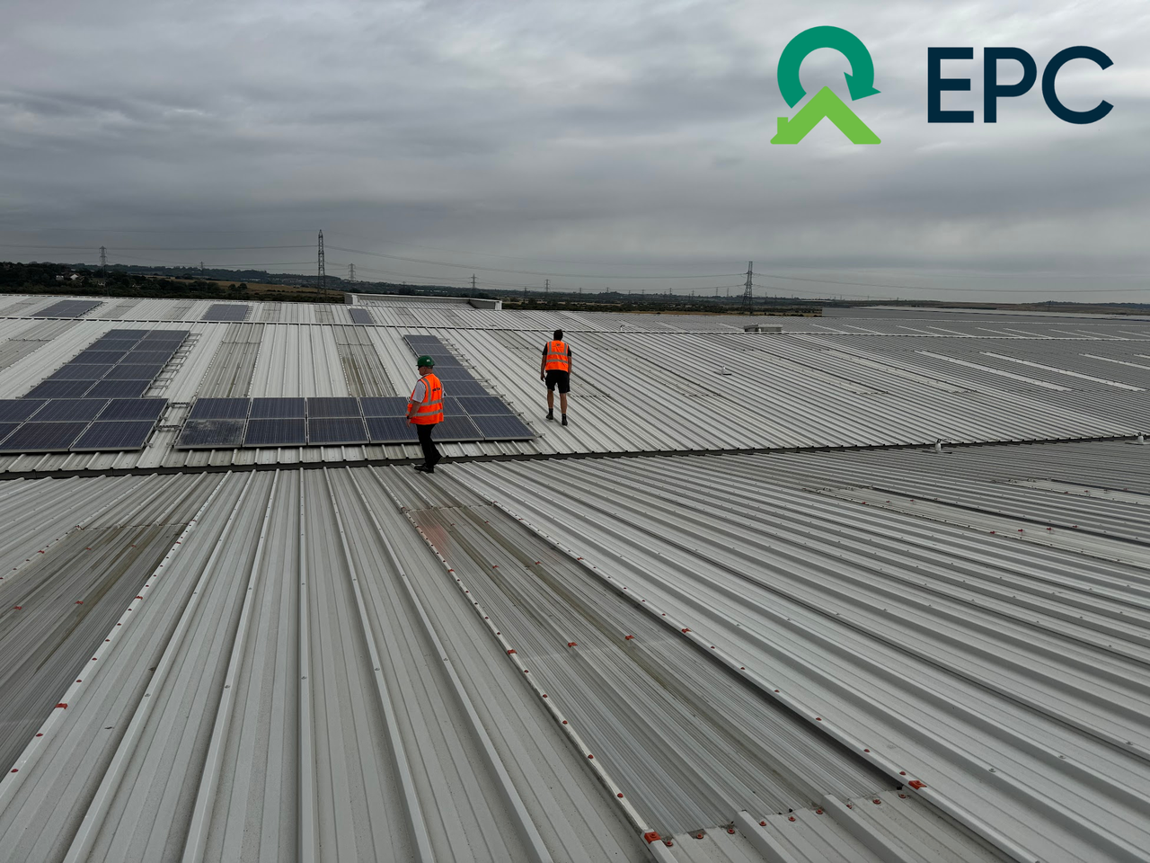 Two workers in orange vests install solar panels on a large corrugated metal roof under a cloudy sky. EPC logo in the top right corner.