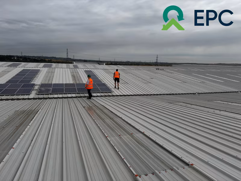 Two workers in orange vests install solar panels on a large corrugated metal roof under a cloudy sky. EPC logo in the top right corner.