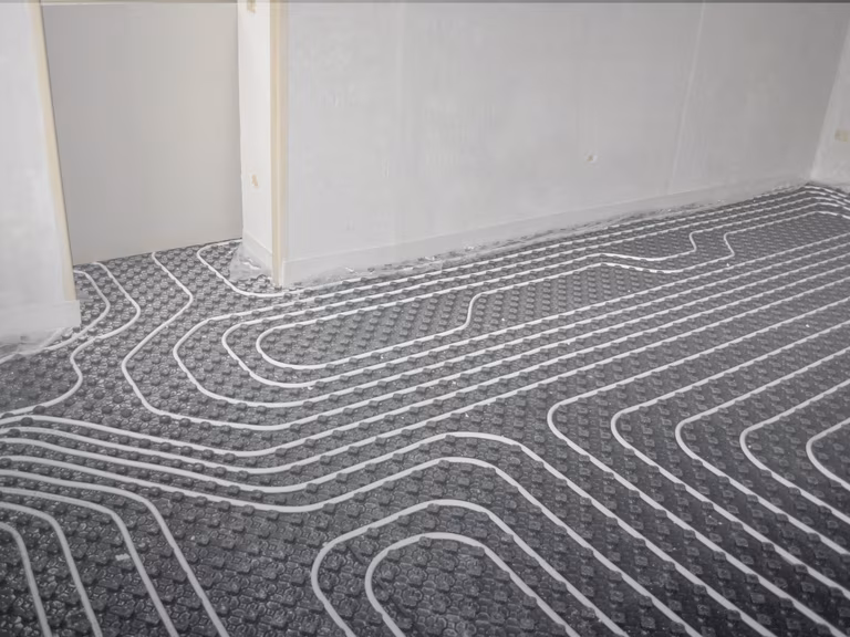 Underfloor heating system installation with visible coiled pipes on a textured black surface in a room with unfinished walls.
