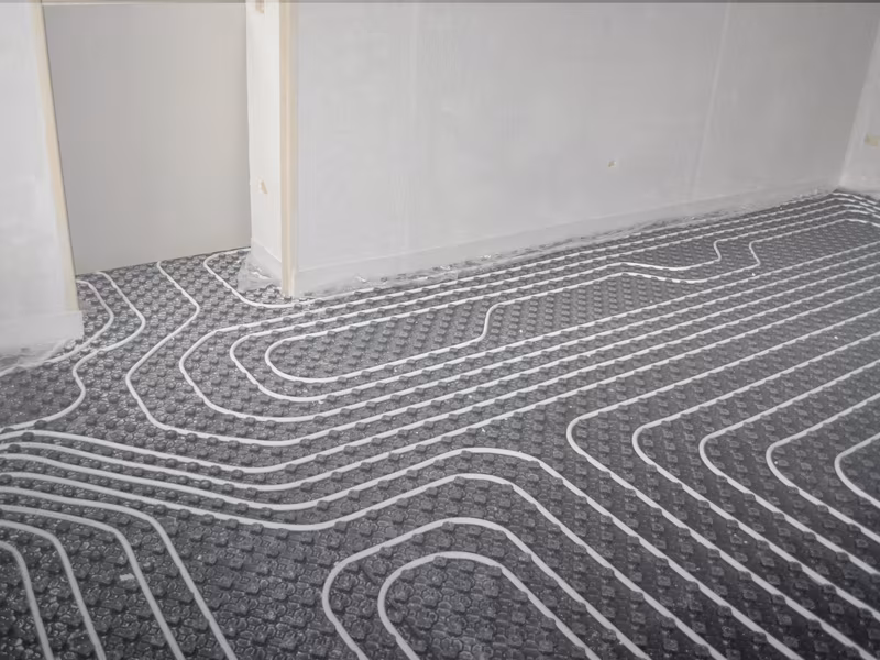 Underfloor heating system installation with visible coiled pipes on a textured black surface in a room with unfinished walls.